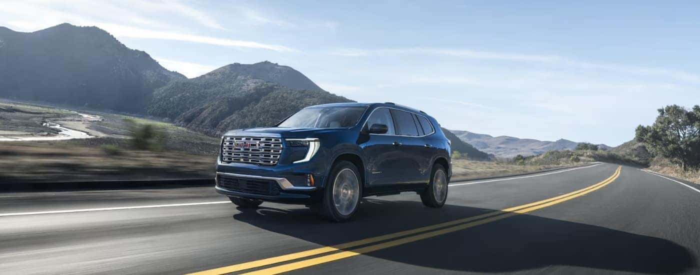 A blue 2024 GMC Acadia for sale driving past a river.