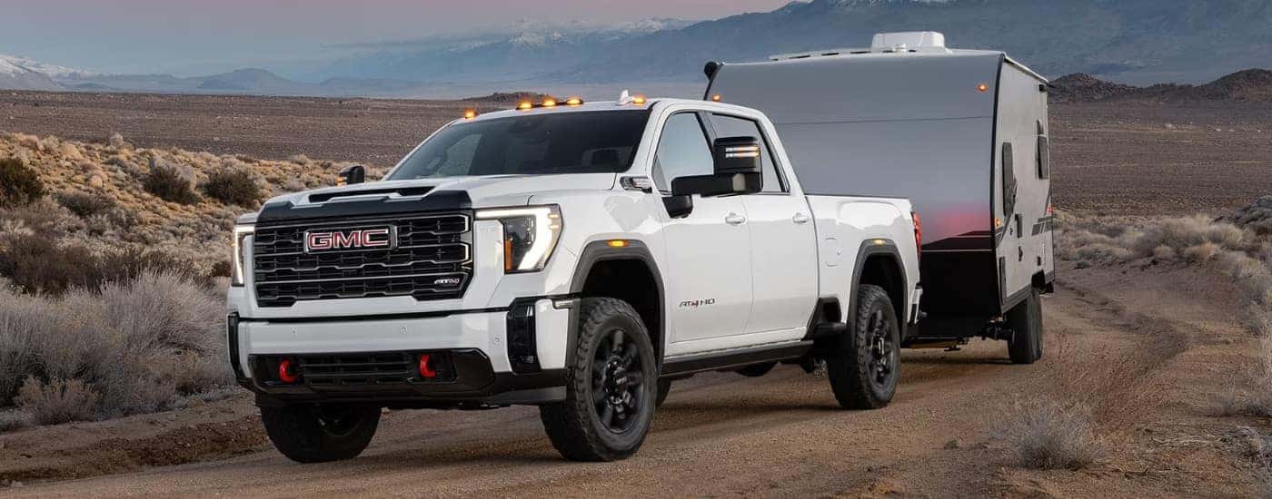 A white 2025 GMC Sierra 2500 HD AT4 is shown towing a trailer off-road.