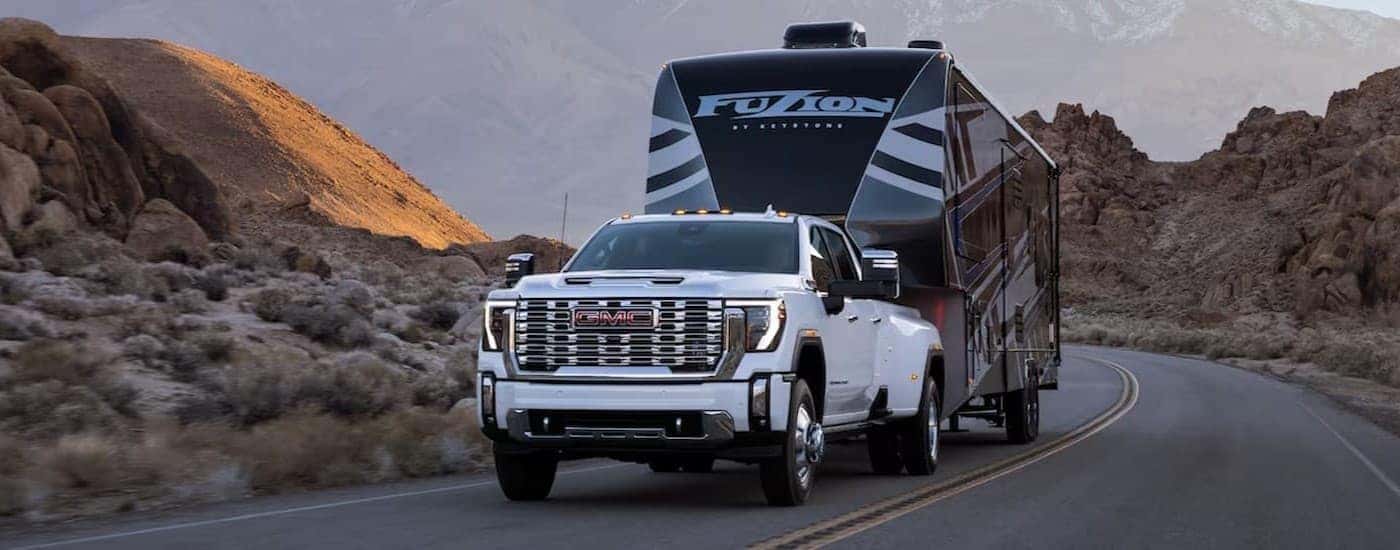 A white 2024 GMC Sierra 2500 HD Denali towing a trailer on a highway.