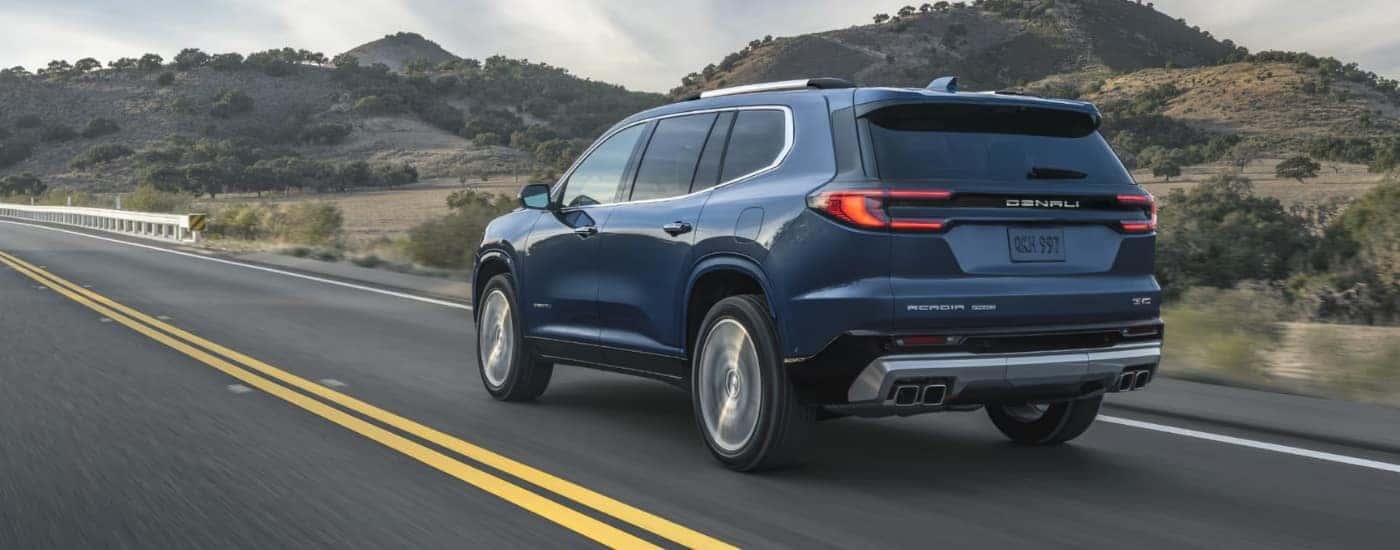 Rear view of a blue 2025 GMC Acadia Denali driving down a highway