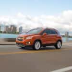 An orange 2015 Chevy Trax driving past water near a city.