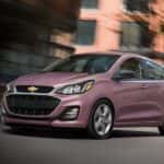 A pink 2021 Chevy Spark is driving past a brick building after leaving a Chevy dealer.