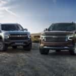 A grey 2021 Chevy Tahoe is next to a brown 2021 Suburban in a rural parking area in front of a blue sky.