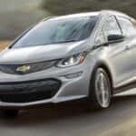 A silver 2021 Chevy Bolt EV is driving passed blurred trees after leaving a Chevy Dealer in Houston.