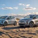 Two silver Chevy EVs, a 2022 Bolt and 2022 Bolt EUV, are parked on beach sand.