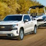 A white 2021 Chevy Silverado 1500, popular among Chevy trucks, is towing a boat past trees.