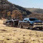 A black 2020 Chevy Silverado 1500 is shown parked near a trail towing two UTVs.