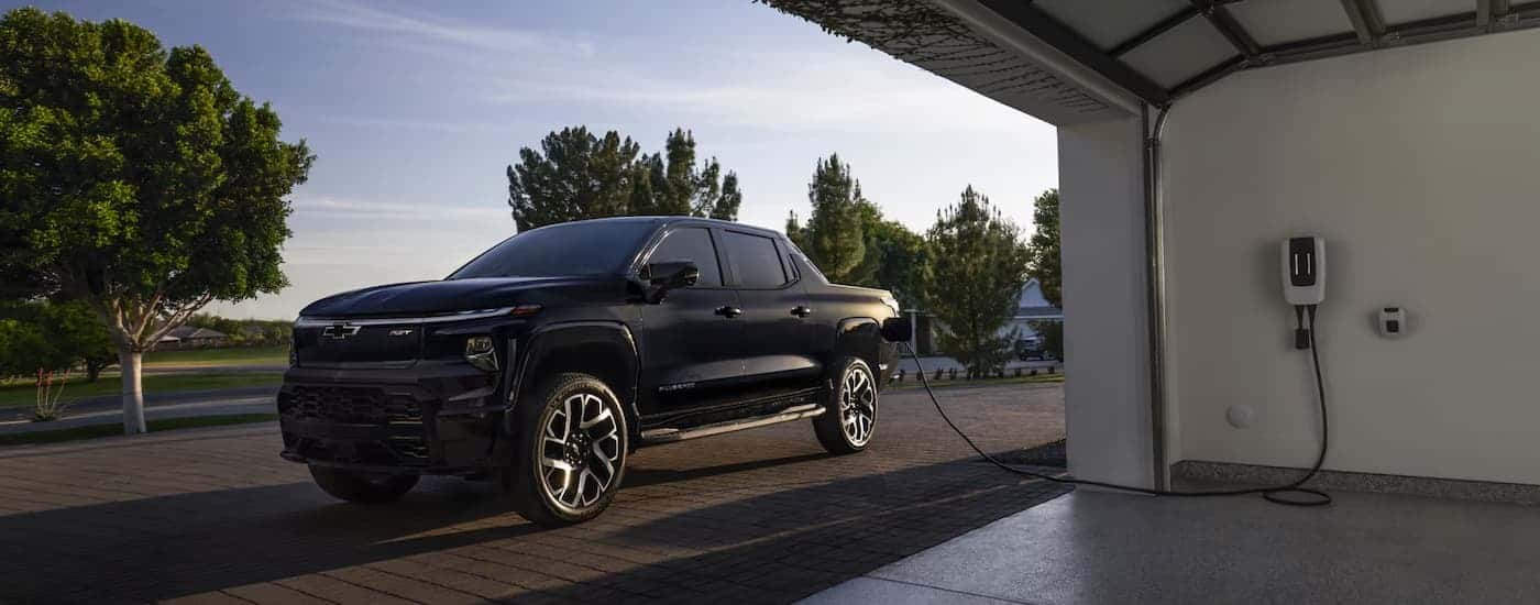 A black 2025 Chevy Silverado EV is charging in a driveway outside of a garage