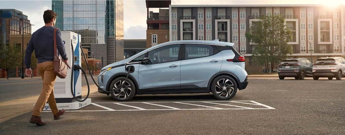 Man approaching a light blue 2022 Chevy Bolt EV at a charging station after leaving a Pinehurst Chevy dealer.