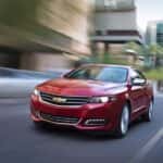 A red 2016 Chevy Impala is driving on a city street after leaving a used car dealer.
