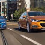 An orange and a blue 2017 Chevy Cruze are driving on a city street with multiple lanes.