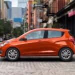 An orange 2021 used Chevrolet Spark is shown from the side driving on a city street.