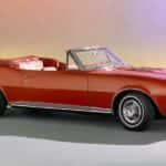 A red 1967 Chevy Camaro convertible is parked in a showroom with a multi-color background.
