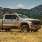 A silver 2019 Chevy Silverado 1500 is covered in mud in a field in front of hills.