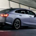 A silver 2019 Chevy Malibu is shown from the rear parked next to a building after visiting a used car dealer.