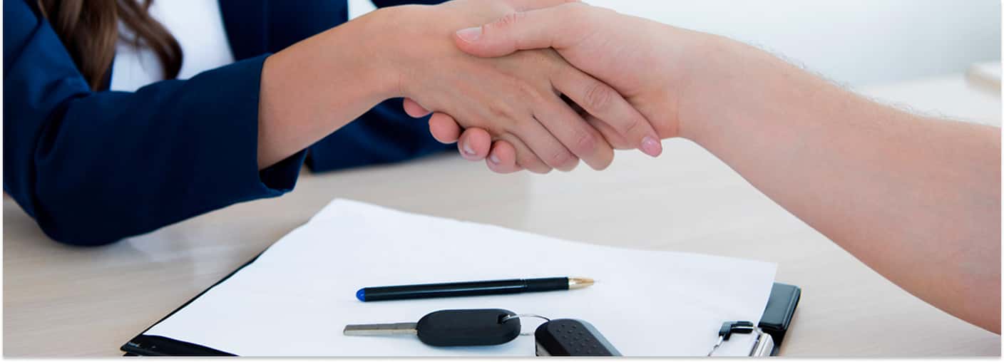 Two people shaking hands across a desk with car keys, a pen, and contract paperwork on a clipboard.