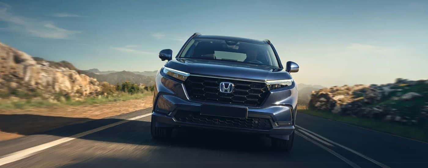 Front of a blue 2026 Honda CR-V Hybrid driving down a road.