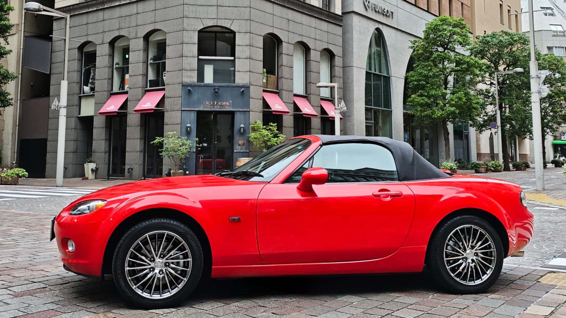 Red Mazda MX-5 Miata convertible side profile with black soft top, chrome wheels, and city street setting.