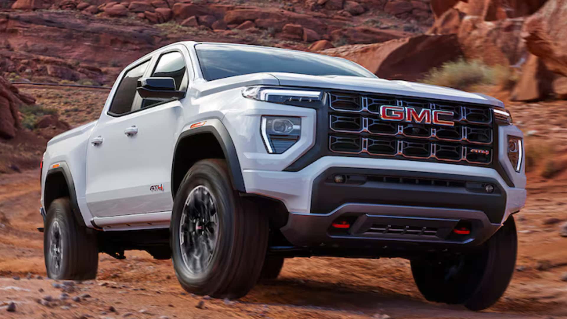 White GMC Canyon AT4X off-road pickup driving through red rock terrain with rugged tires and lifted stance.