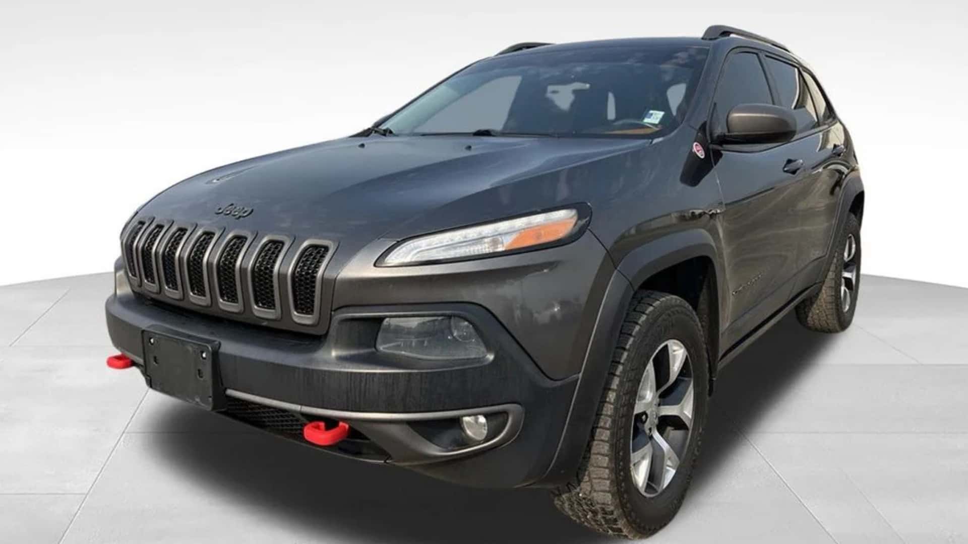 Dark gray Jeep Cherokee front three-quarter view showing seven-slot grille, red tow hooks, roof rails, and rugged tires.