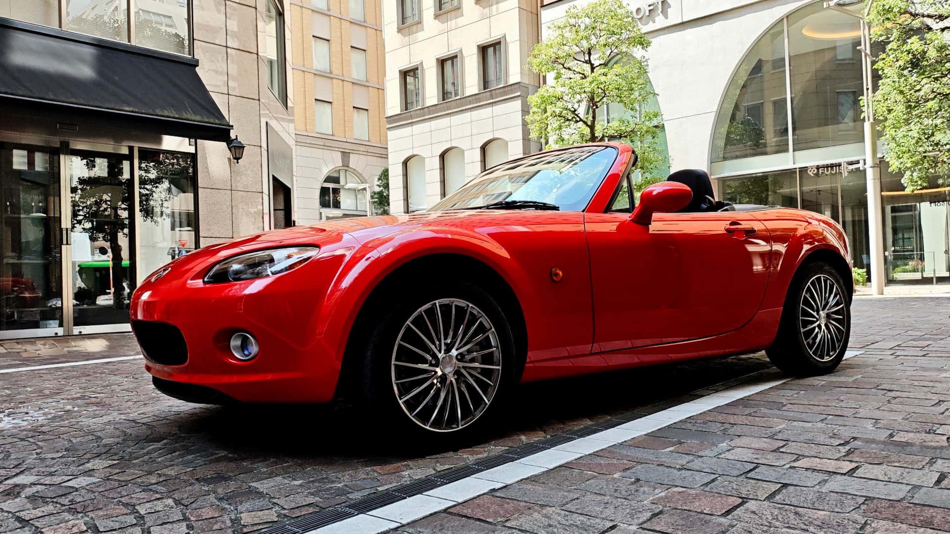 Red Mazda MX-5 Miata convertible parked on a city street with black soft top down and sporty roadster styling.