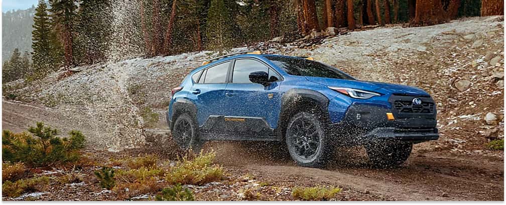 Front three-quarter view of a Geyser Blue 2026 Subaru Crosstrek Wilderness splashing mud on a snow-lined trail.