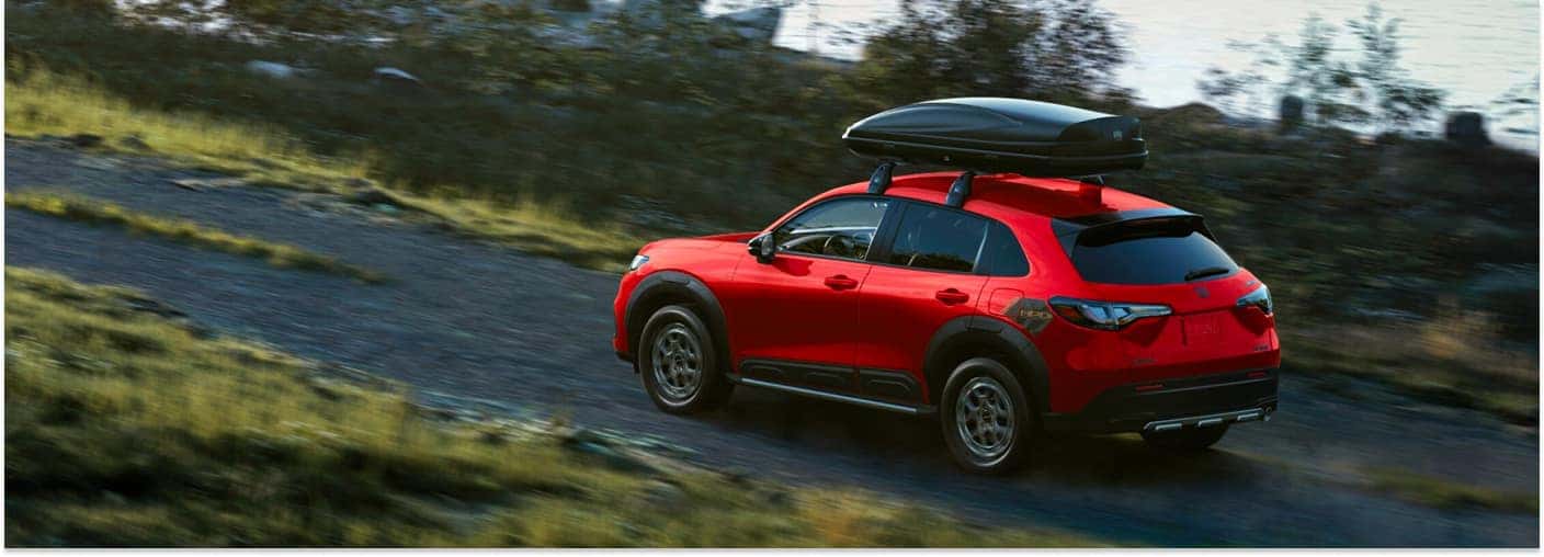 Rear three-quarter view of a red 2026 Honda HR-V driving on a dirt road with a roof cargo box.