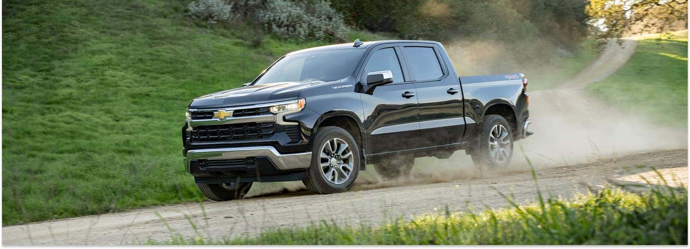 A black 2026 Chevrolet Silverado 1500 pickup truck kicks up dust while driving along a dirt trail, viewed from a front three-quarter angle with a background of rolling green hills.