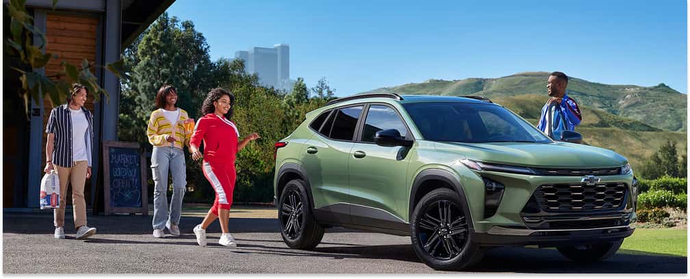 A green 2026 Chevrolet Trax compact SUV is parked outside a market building on a sunny day, with a group of friends interacting around the vehicle against a backdrop of rolling green hills and a distant city skyline.