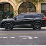 A black 2026 Subaru Ascent is parked along a city street, showcasing its side profile and black wheels against a storefront background.