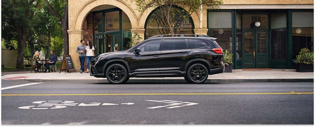 A black 2026 Subaru Ascent is parked along a city street, showcasing its side profile and black wheels against a storefront background.