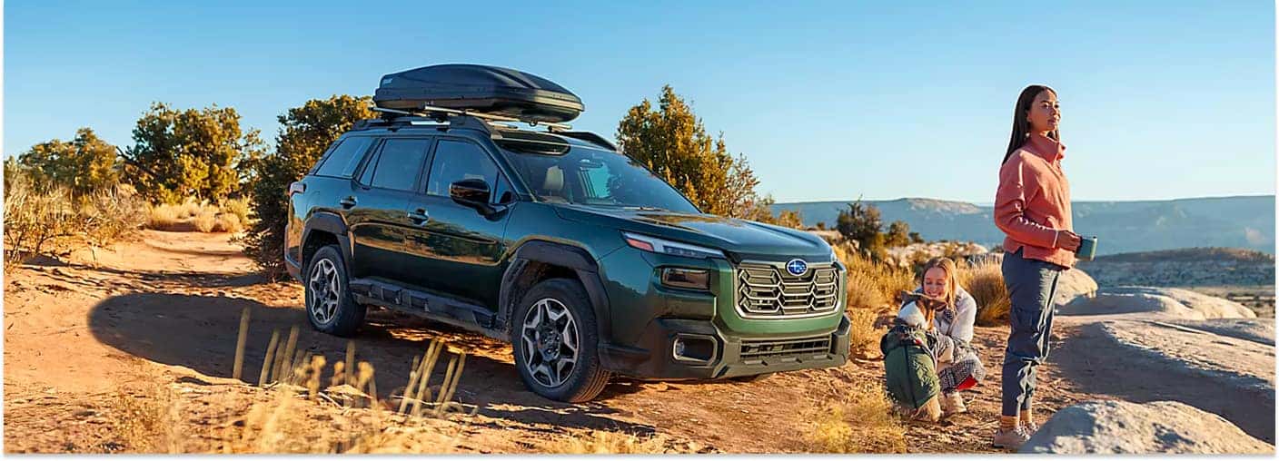 Dark green 2026 Subaru Outback parked on a dirt trail, front three-quarter view with a roof cargo carrier.