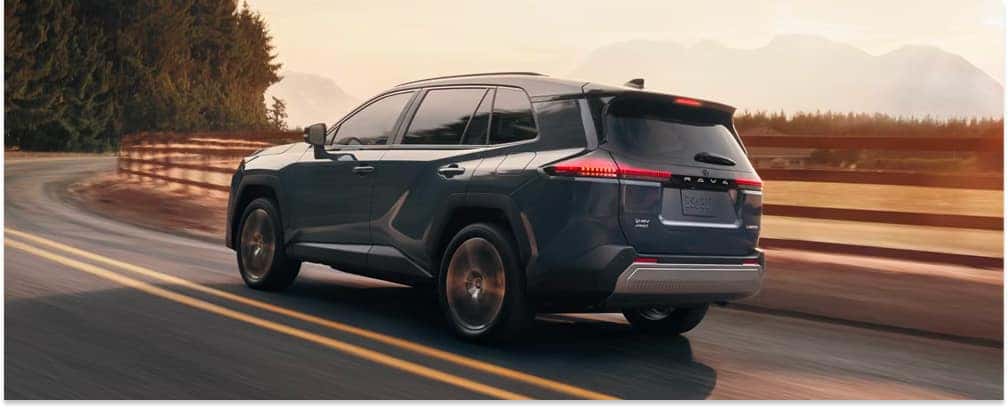 Rear three-quarter view of a dark gray 2026 Toyota RAV4 driving on a mountain road at sunset.