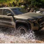 Front three-quarter view of a green 2026 Toyota Tacoma pickup truck splashing through a rocky stream.