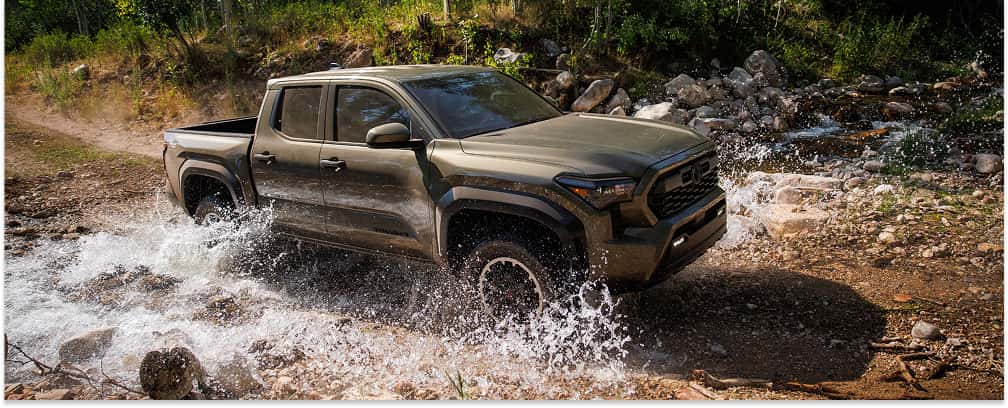 Front three-quarter view of a green 2026 Toyota Tacoma pickup truck splashing through a rocky stream.