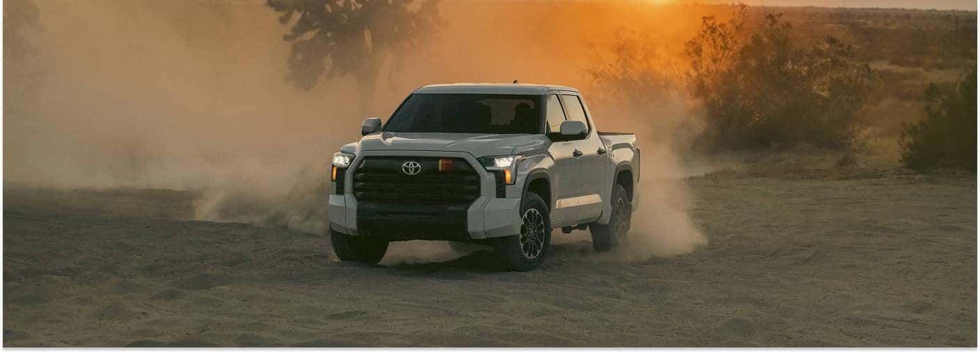 Front three-quarter view of a white 2026 Toyota Tundra driving on a dusty desert terrain at sunset.