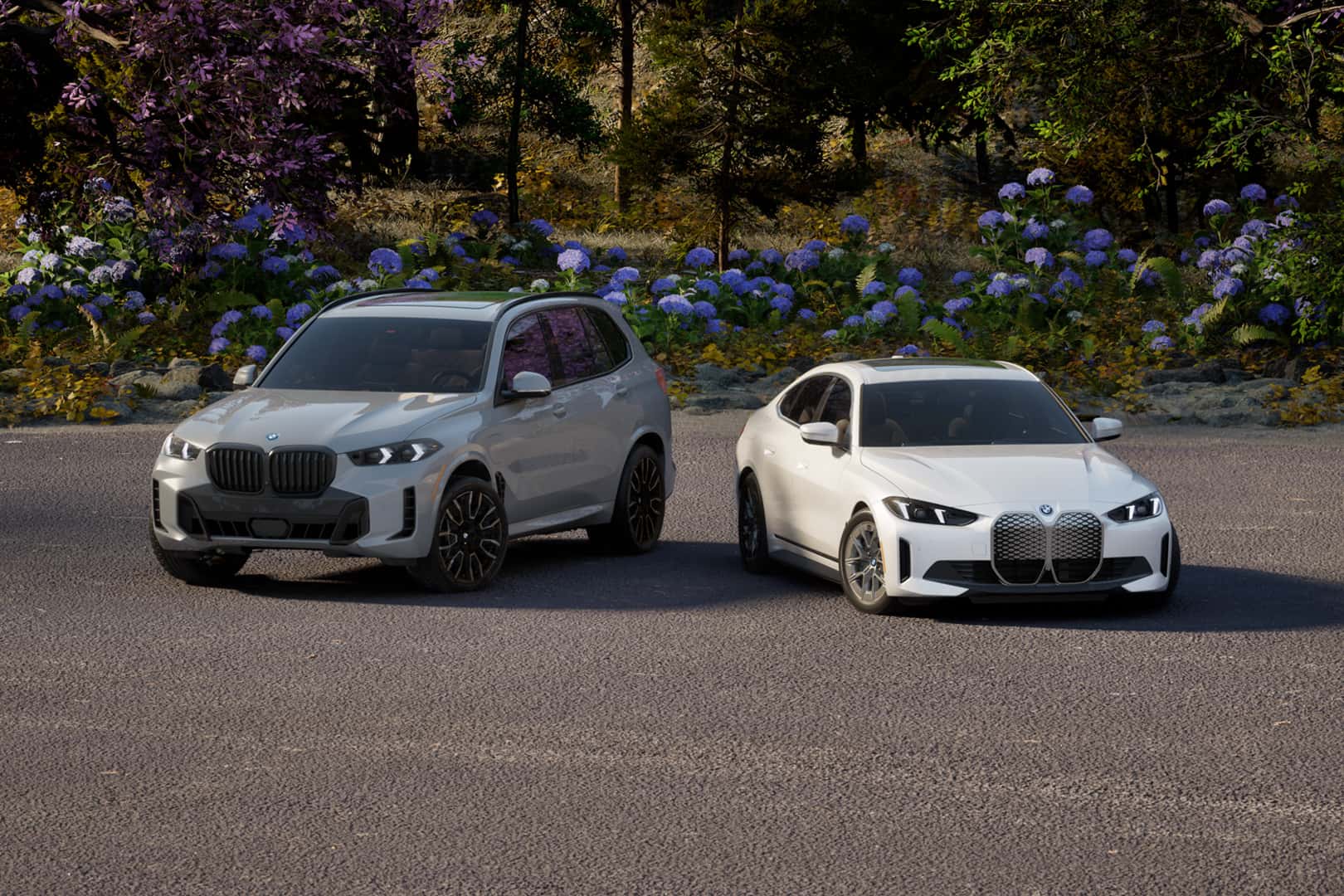 White BMW SUV and white BMW sedan parked side by side on a paved road, with colorful flowers and trees in the background in a natural outdoor setting.