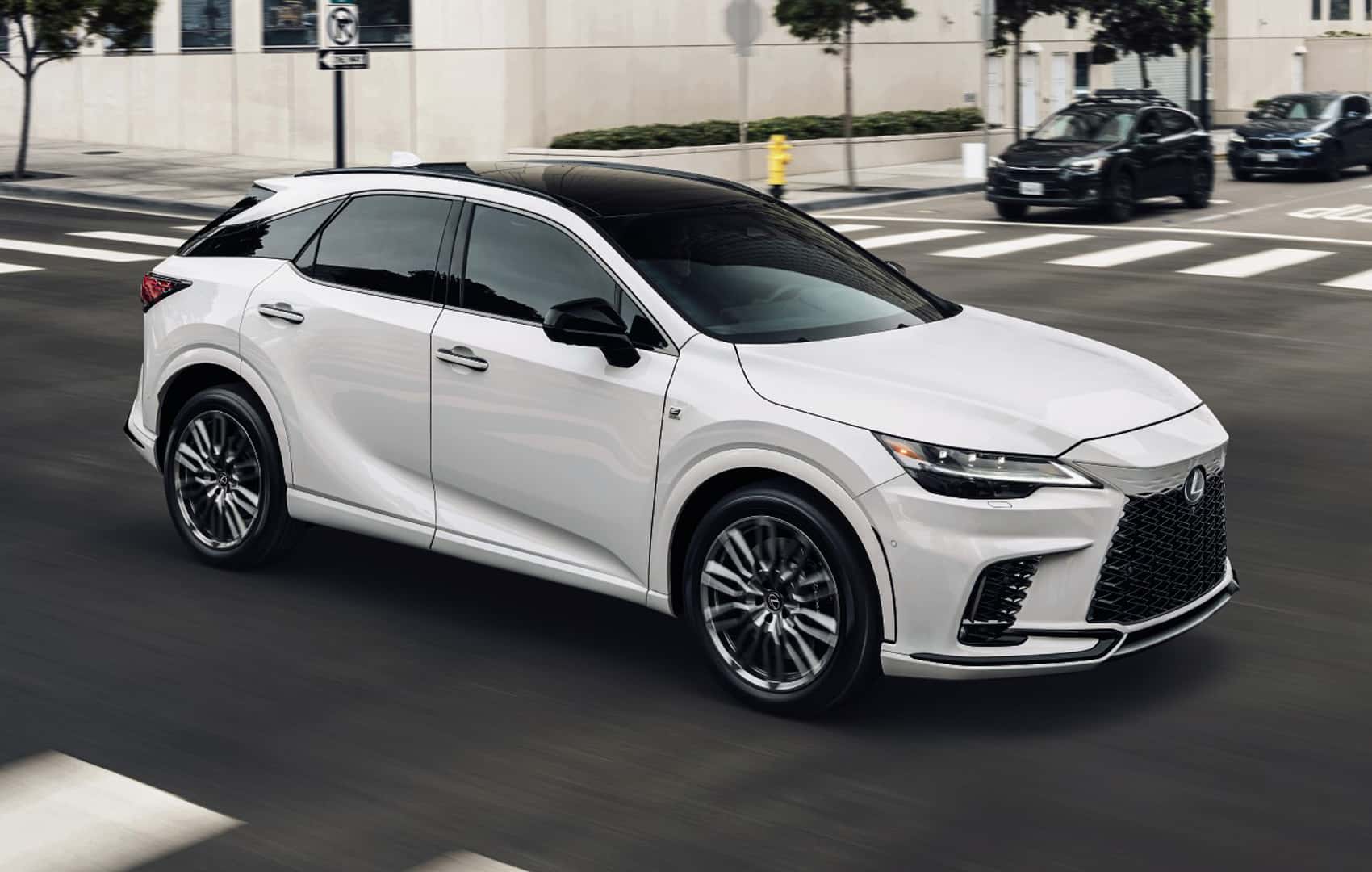 White Lexus RX SUV driving through a city street, featuring a sleek design, black roof, and signature spindle grille.