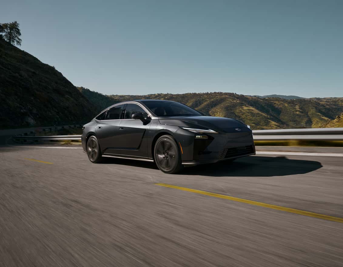 Dark gray Lexus ES sedan driving along a winding mountain road, featuring a sleek silhouette, modern front fascia, and scenic hillside landscape in the background.