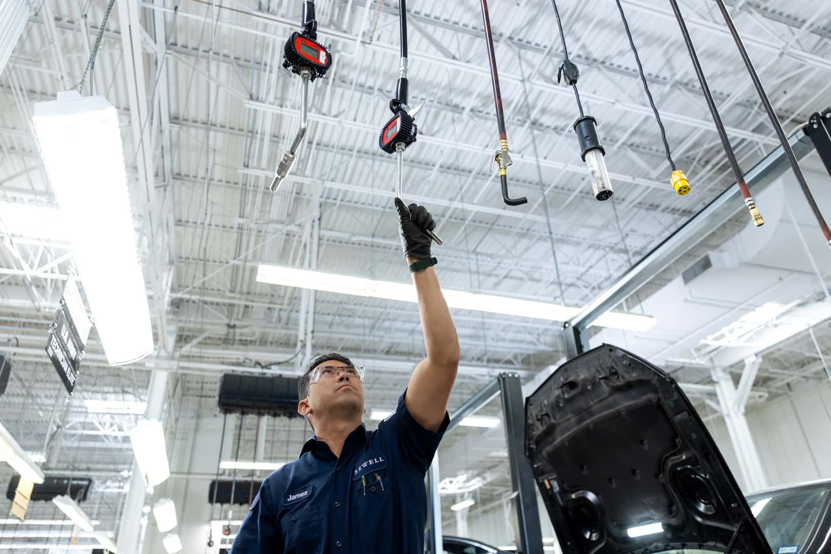 Sewell technician in a Sewell service bay reaches up to secure a suspended air tool hose while working near an open vehicle hood in a bright, modern automotive workshop.