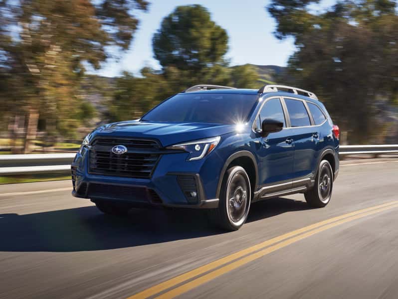 Blue Subaru Ascent SUV driving on a paved road with trees and hills in the background under bright daylight.