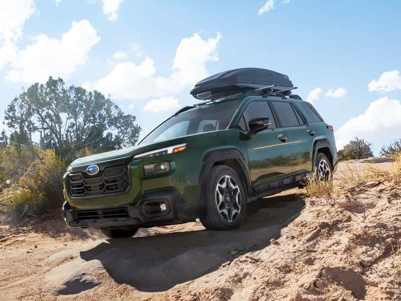 Green Subaru Outback with roof cargo box driving down a sandy off road trail, surrounded by desert brush under a clear blue sky.