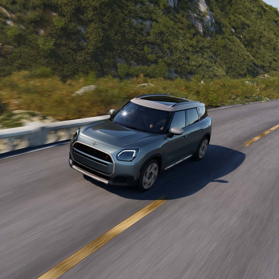 Green MINI Countryman driving on highway with grassy hill on the shoulder.