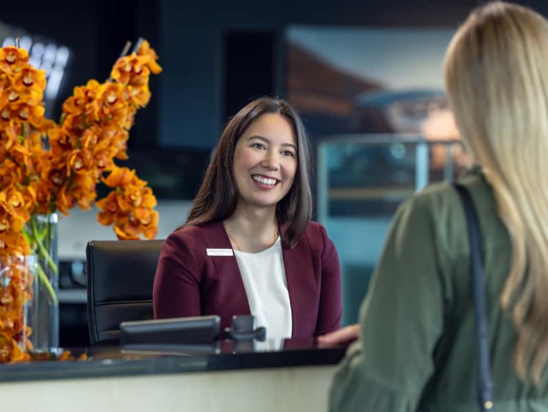A Sewell business admin associate smiles while assisting a guest at a store reception desk. Orange floral arrangements accent the welcoming showroom environment, reflecting a professional and customer focused experience.