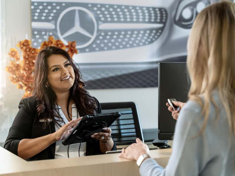 A Sewell business admin associate assists a guest at a dealership reception desk while reviewing information on a tablet. A Mercedes-Benz display is visible in the background, highlighting a welcoming and professional customer experience.
