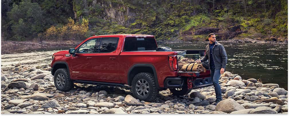 Rear three-quarter view of a red 2026 GMC Sierra 1500 parked on a rocky riverbank with its tailgate open.