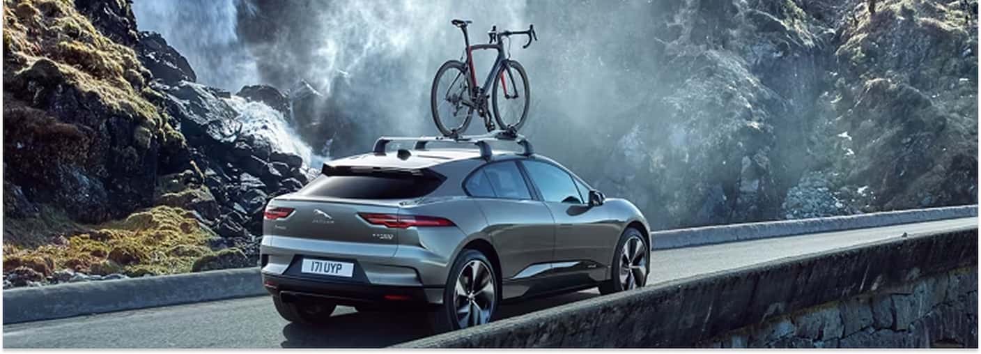 Rear three-quarter view of a grey Jaguar I-PACE equipped with a roof rack and bicycle near a waterfall.