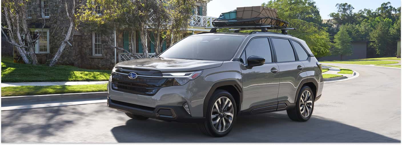 Front three-quarter view of a grey 2026 Subaru Forester with a loaded roof rack driving on a residential street.