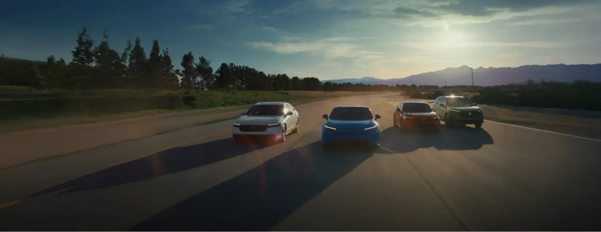 Four new Honda vehicles, including a sedan, the Prologue EV, and two SUVs, driving side by side down an open road at sunset