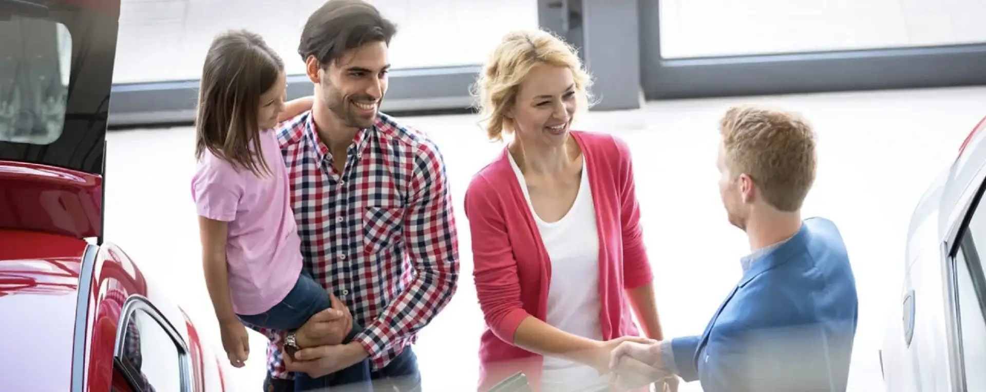 A smiling family with a young daughter shakes hands with a salesperson at a car dealership showroom